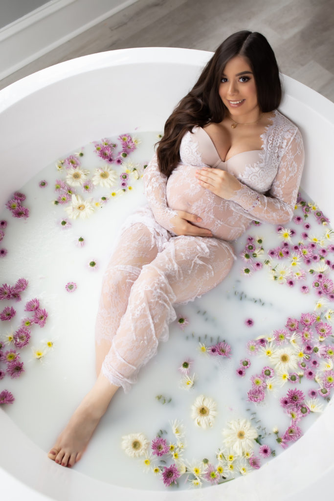 Pregnant mother in birthing tub filled with milk and flowers wearing lace dress for maternity photo session getting pictures done in birthing suite at Origins Birth Dallas