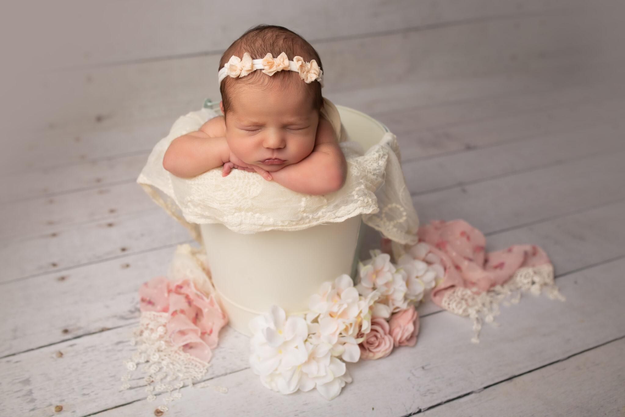 Dallas newborn photographer poses baby girl in cream colored bucket with chin on hands surrounded by flowers and lace scarf for texture