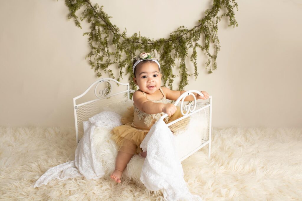 Dallas baby photographer poses 9 month old girl on mini bed wearing a gold tutu she is smiling