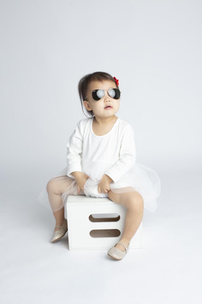 Dallas child photographer shows portrait of young girl seated on white box wearing white dress and sunglasses.