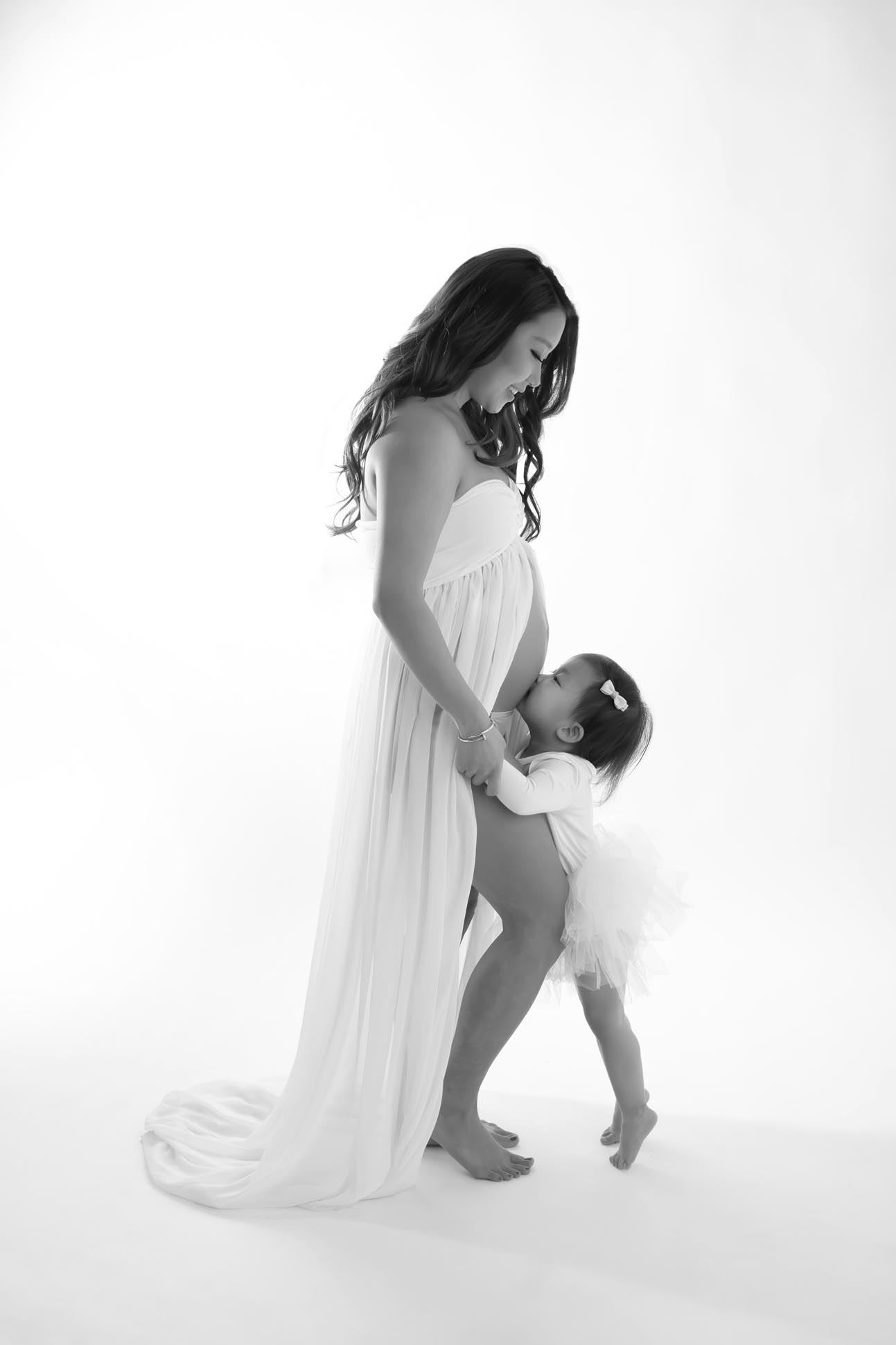 Dallas maternity photographer shares portrait of mom to be with big sister sibling kissing belly both are wearing all white dresses