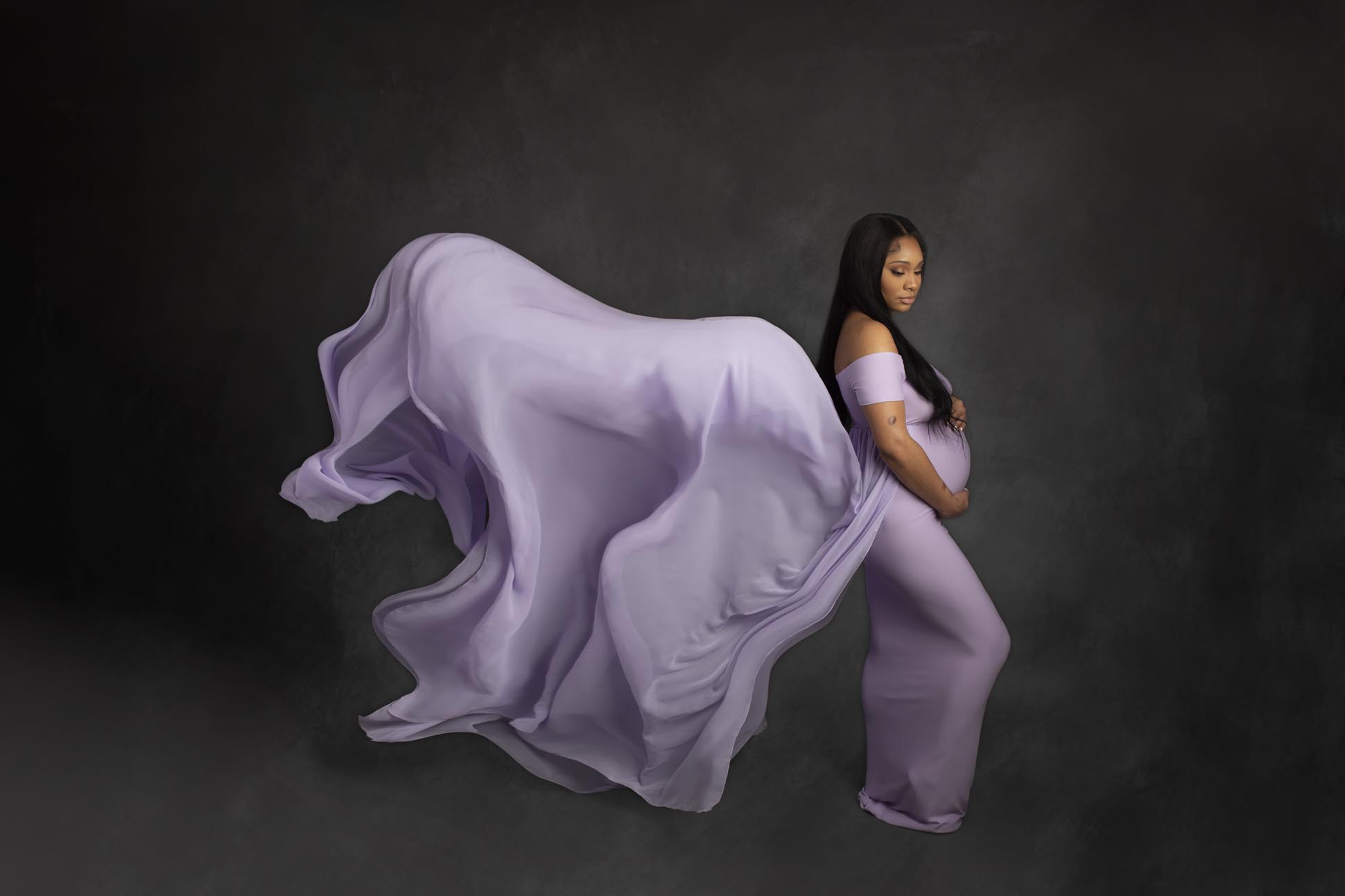 Dallas maternity photographer poses pregnant mom to be in lavender dress with flowing train on dark gray backdrop during studio portrait shoot