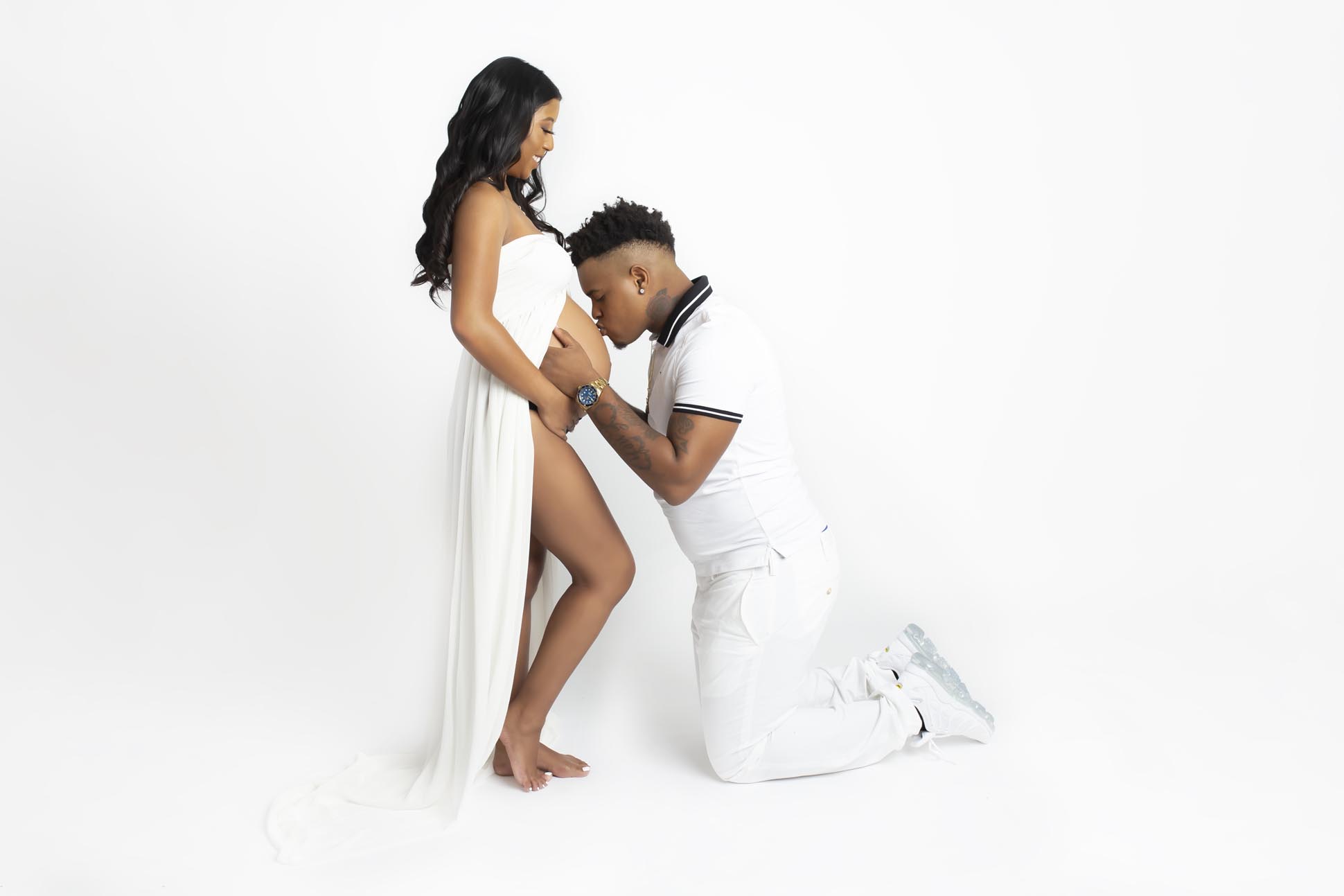 Dallas maternity photographer shares portrait of parents to be wearing all white on a white background dad is kneeling and kissing mom's bared body
