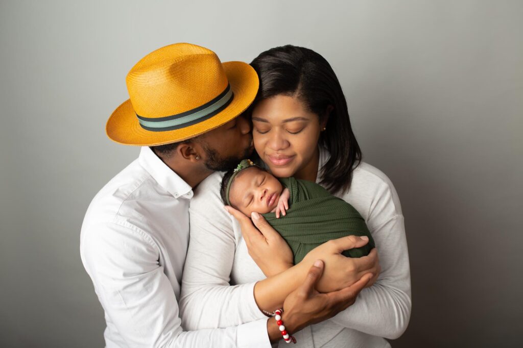 How to pose newborns with pare holding baby girl swaddled in green dad is wearing golden yellow fedora and leaning over to kiss mom on the cheek