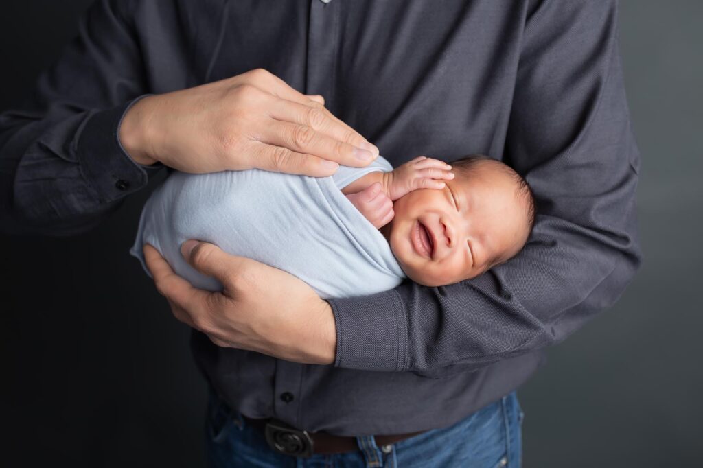 dallas newborn photographer poses baby like a football in his dad's arms, baby is swaddled and dad holds him in his arm just like a football