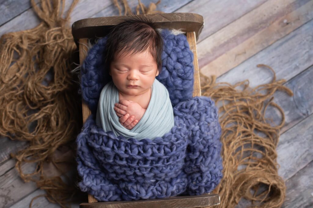 Dallas newborn photographer poses baby boy in blue wrap and darker blue knit blanket in a rustic wooden bed on a blue toned wood floor during his baby photography session