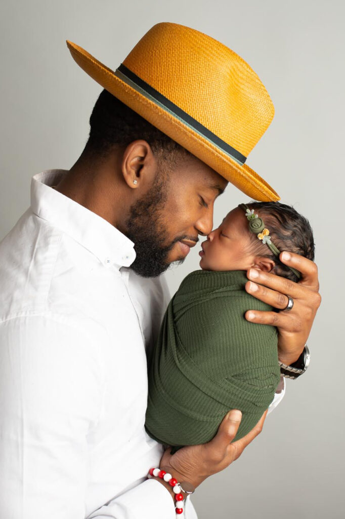 Dallas maternity newborn portrait photographer captures portrait of new dad with baby daughter. Dad is wearing white shirt and golden yellow fedora, daughter is wearing green swaddle wrap and matching headband. They are pictured nose to nose.