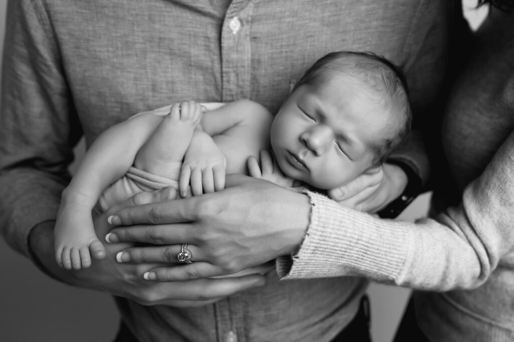 Newborn photographer poses baby in dads arms and has mom come in with her hand over baby protectively showing a great how to pose newborns with parents