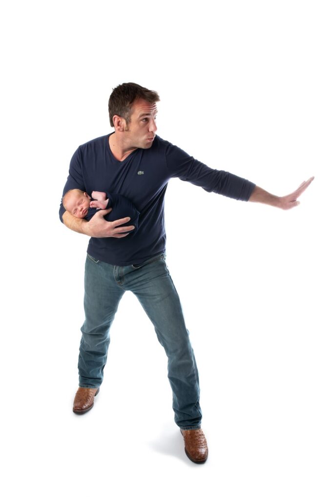 New dad pictured holding his infant baby boy like a football in classic football pose dad is wearing jeans and dark blue shirt on white background during Richardson Texas newborn photography session.