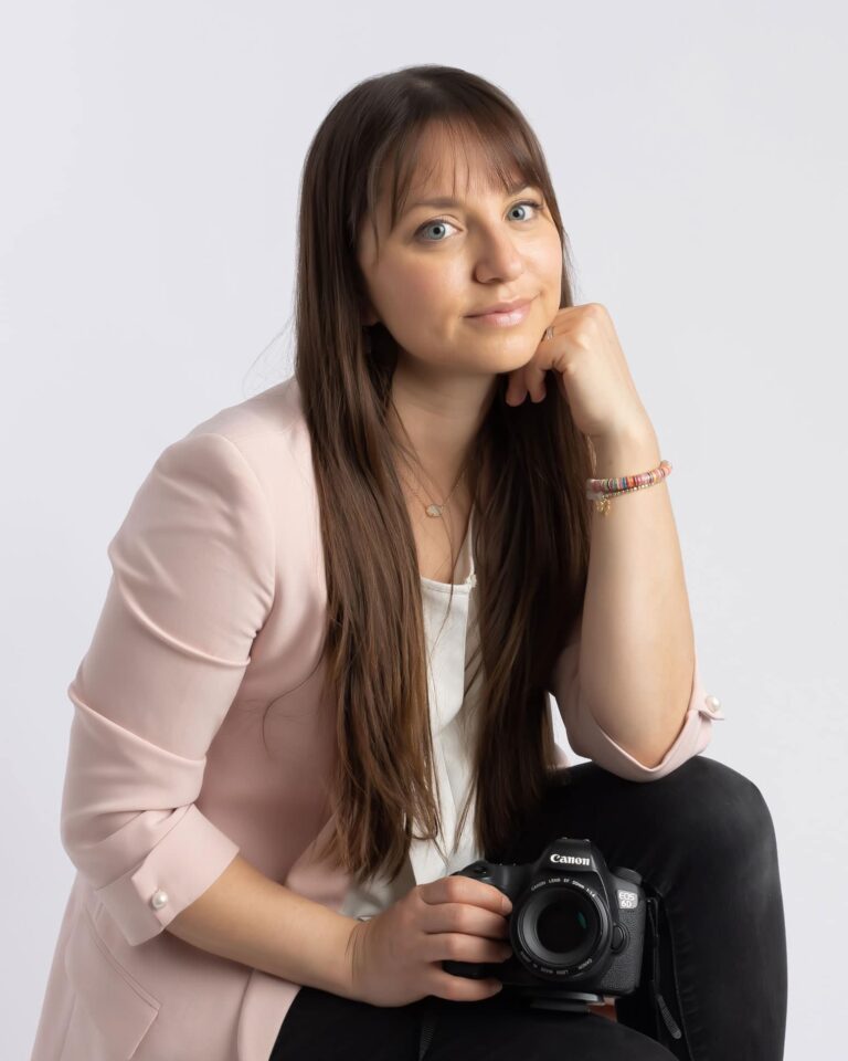 Professional Dallas Photographer Laura Levitan pictured seated with her camera wearing a pink blazer, black jeans and white blouse.