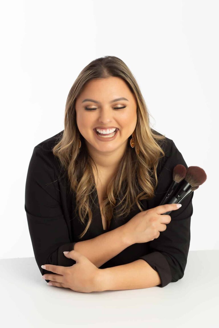 Dallas hair and makeup artist poses for her branding headshot in north dallas photography studio. She is wearing all black and is posed leaning on a table while holding her makeup brushes and laughing.