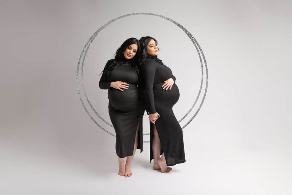 Identical twin pregnancy photoshoot pose next to each other during their maternity session with Mod L Photography a Dallas based maternity and newborn photographer named Laura Levitan, the mothers are twins and wear the same black dress.