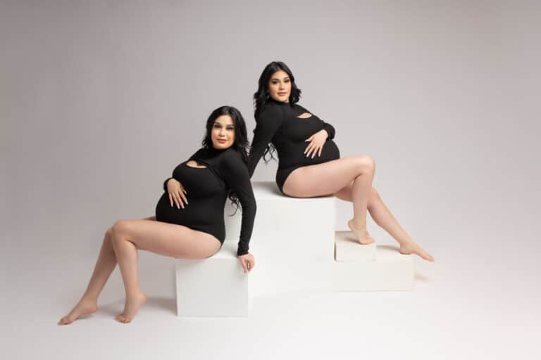 Posing identical twin mothers pregnant at the same time on white posing blocks both wearing black bodysuits by Dallas maternity photographer Mod L Photography - Laura Levitan