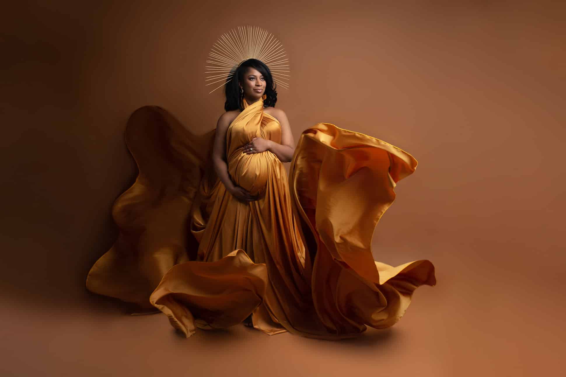 Dallas Maternity photographer poses expectant mom on brown background with flowing gold fabric flying all around and gold crown