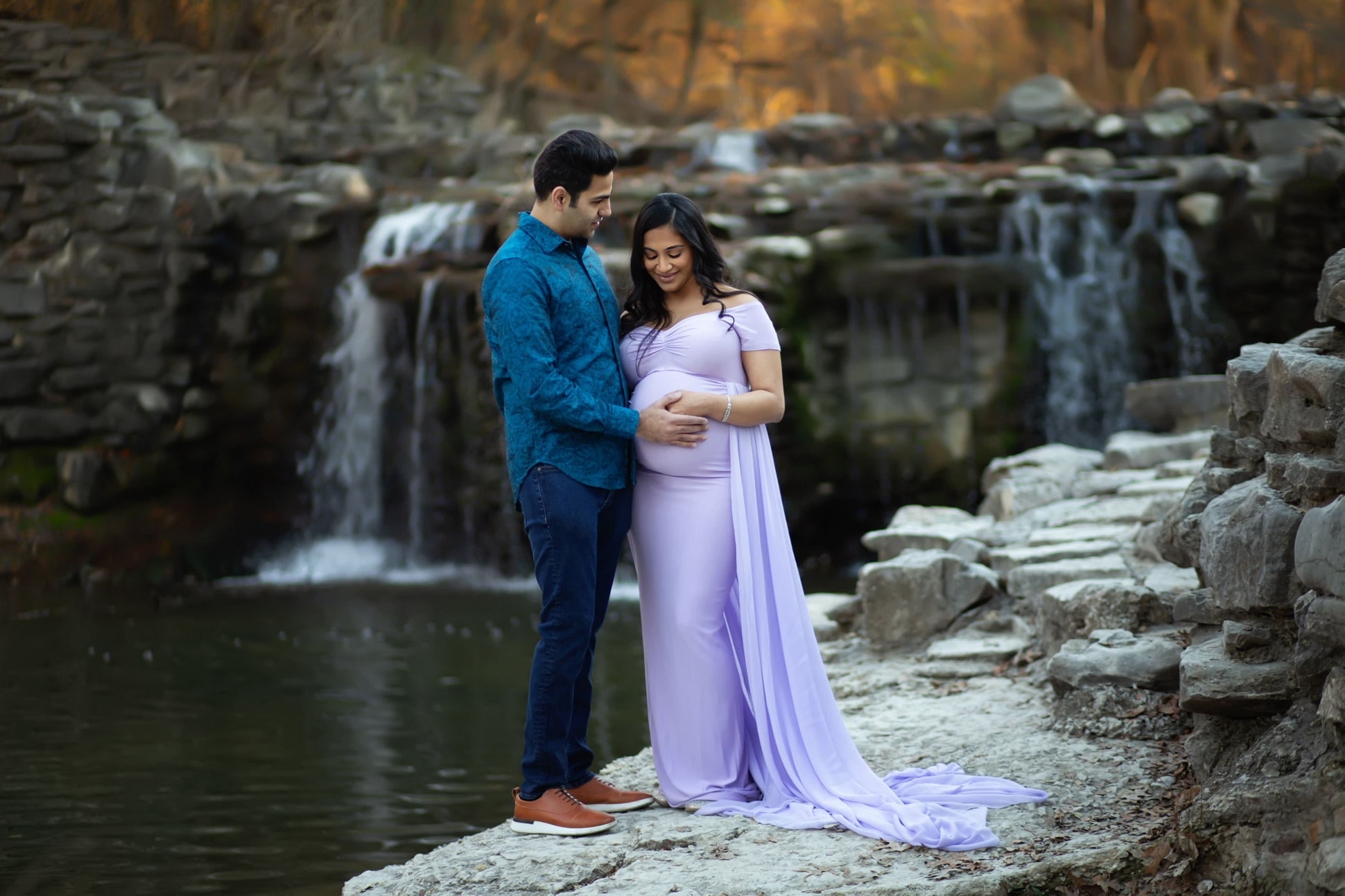 Dallas maternity photographer poses expectant couple in front of Prairie Creek Waterfall in Richardson Texas wearing shades of lavender and blue for their maternity photography session