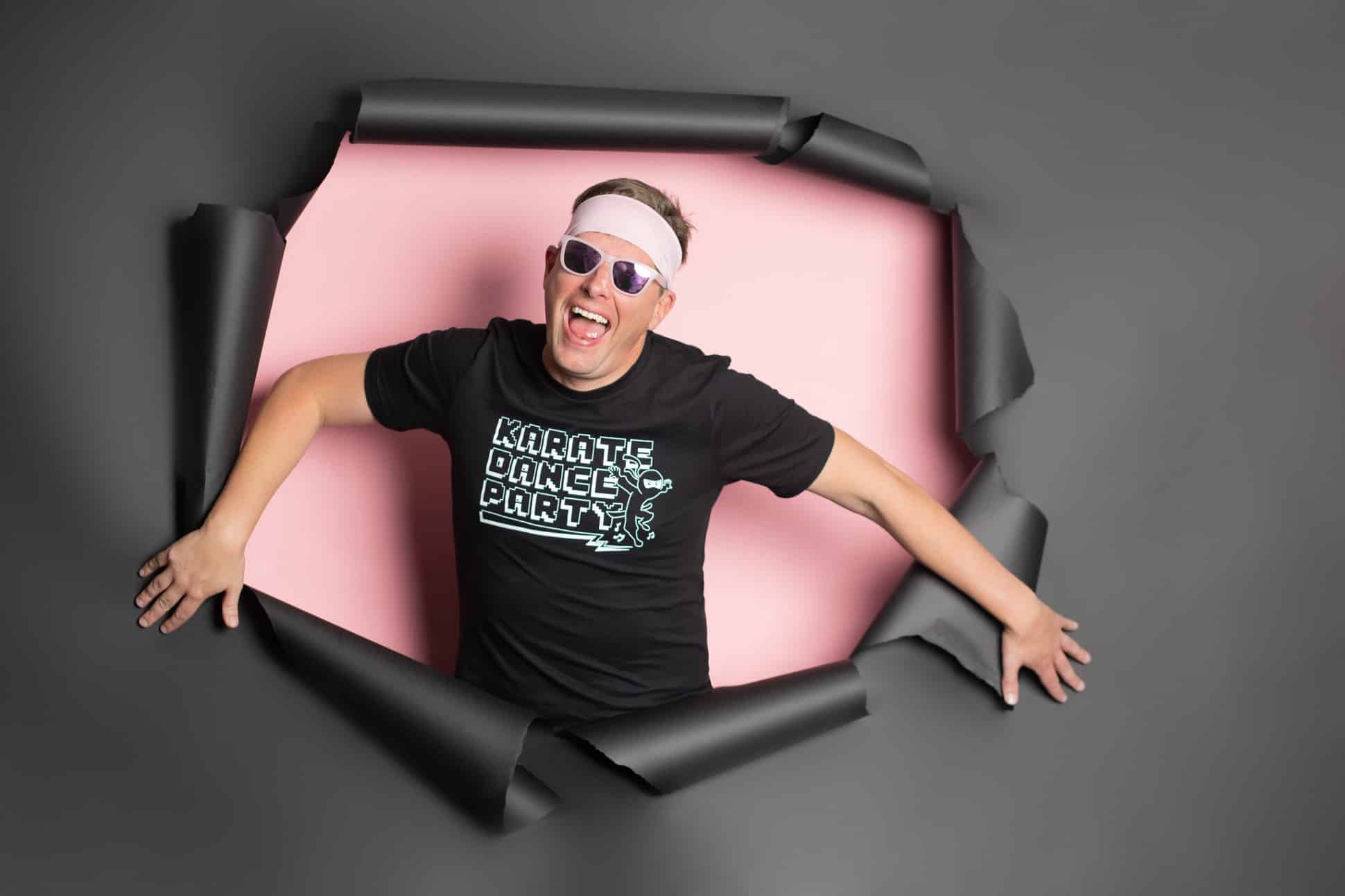 Karate Dance Party owner and lead performer Gene Davis pictured popping out of a black paper backdrop wearing a pink headband and pink sunglasses in his branding headshots by Mod L Photography Dallas photographer