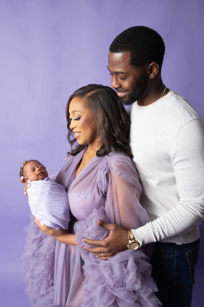 New parents gaze lovingly at their newborn baby girl during photography session baby and mother in lavender on lavender background at Mod L Photography portrait studio in Dallas Texas